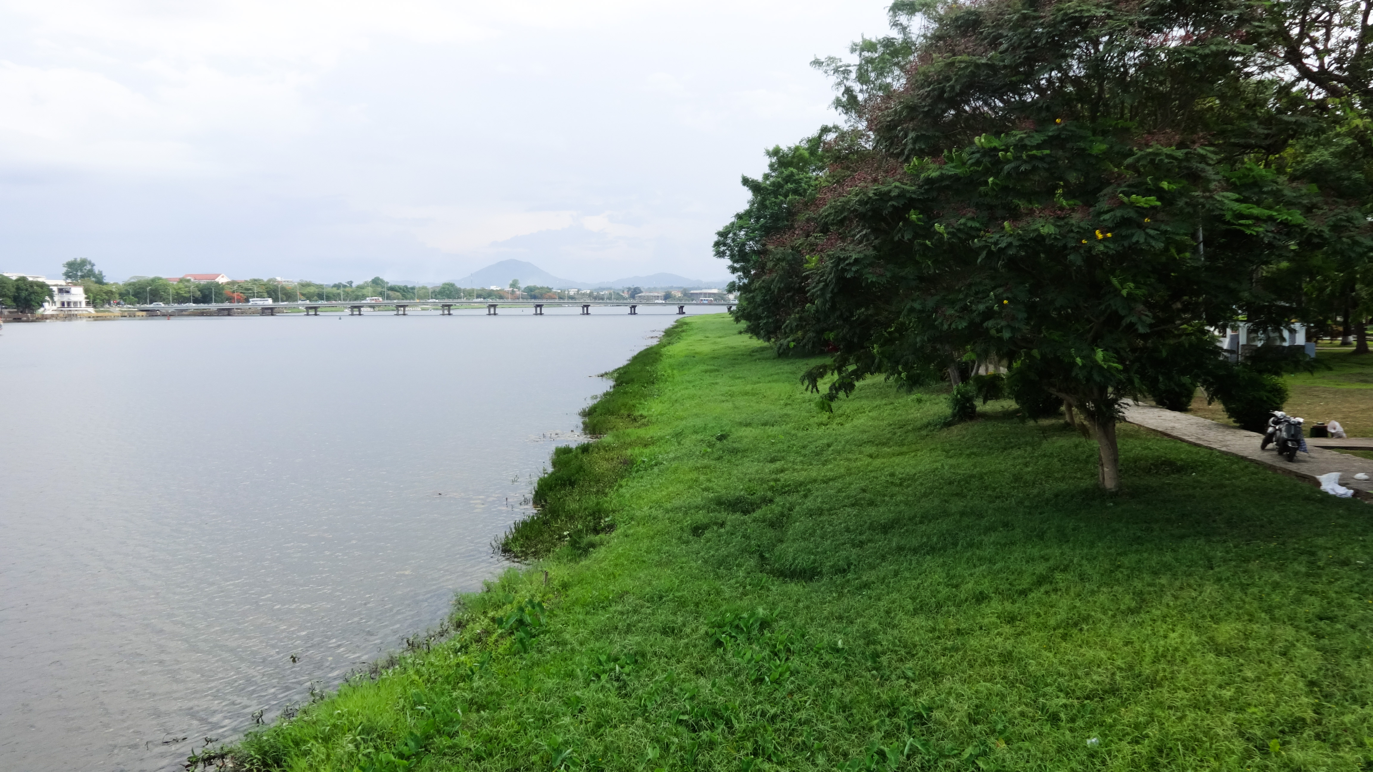 Bờ sông Hương nhìn từ cầu Tràng Tiền — hàng cây rậm rạp và mặt nước yên tĩnh phản chiếu bầu trời Huế